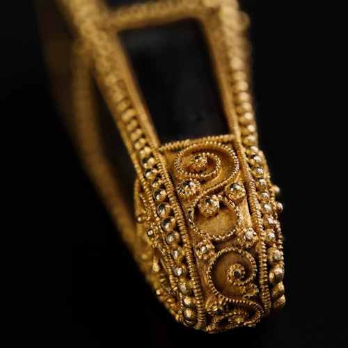 Close-up of an ornate gold bracelet featuring intricate designs and encrusted with small gemstones on a black background.