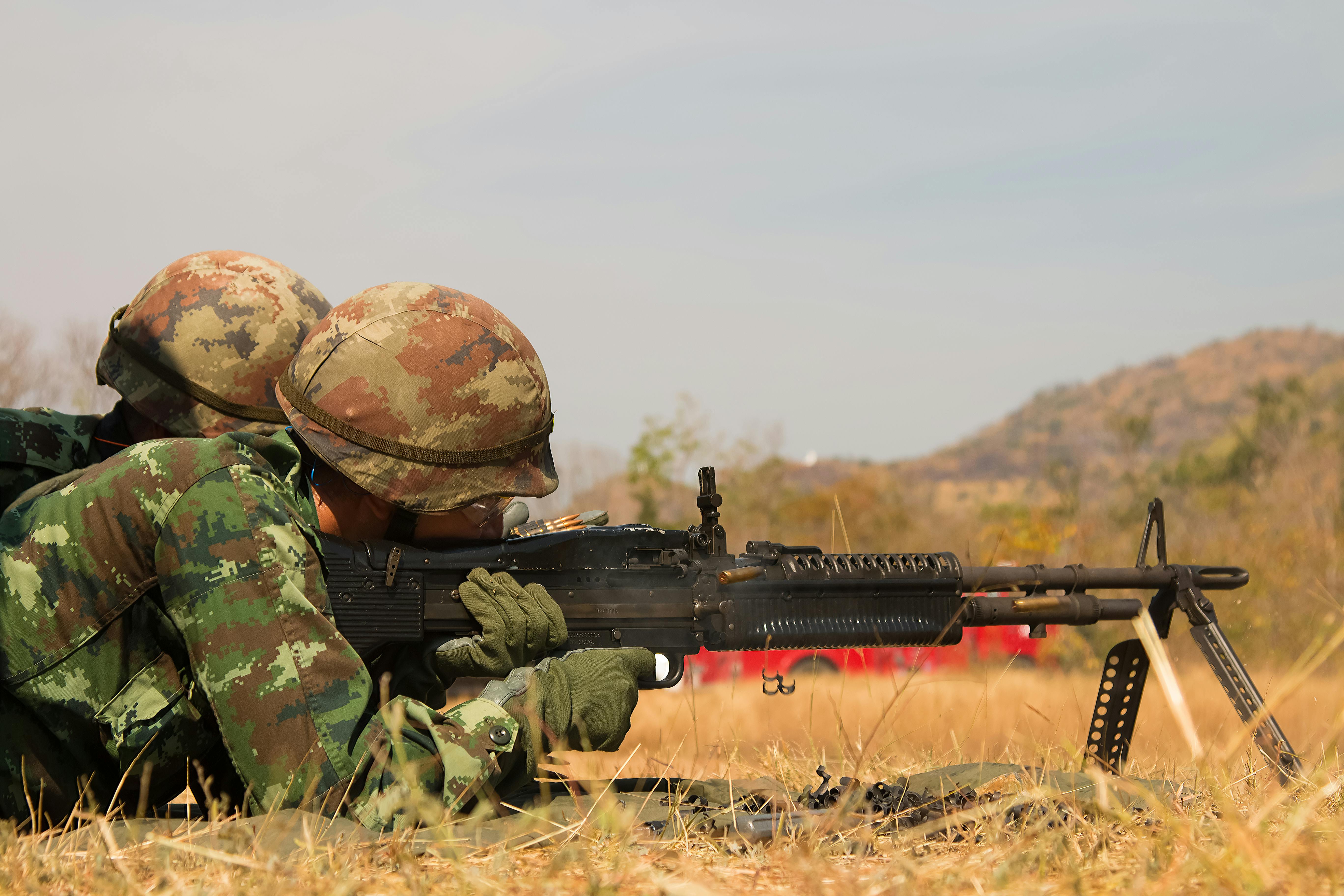 Two soldiers in camouflage uniforms lying prone in dry grass, operating a light machine gun. Both are wearing camouflage helmets with autumn-colored digital pattern. The weapon appears to be mounted on a bipod. In the background is a hilly, arid landscape under a pale sky. The soldiers are positioned in a firing stance during what appears to be a training exercise.
