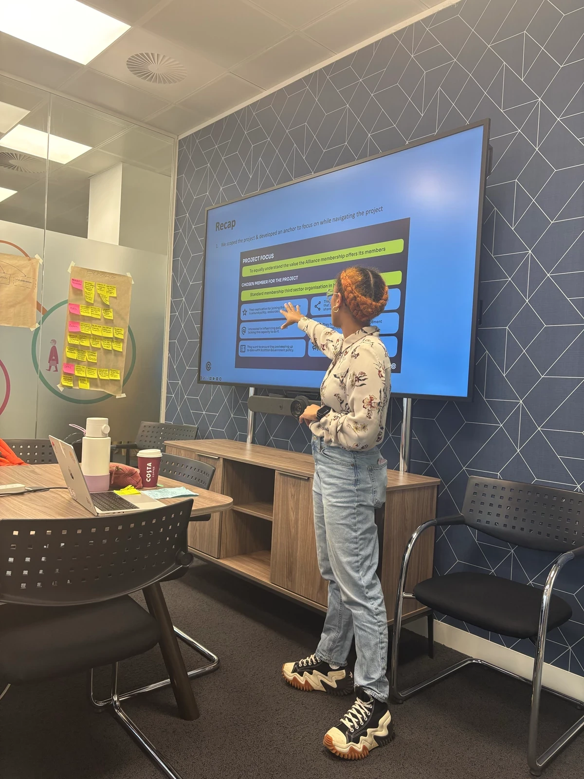 Person standing and pointing at a large screen displaying a presentation slide in a modern meeting room with patterned wall, chairs, and a table with a laptop, coffee cup, and stationery.