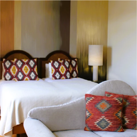 A cozy bedroom featuring a bed with a white quilt and patterned pillows under a warm-toned wall, next to a modern lamp and grey couch with matching cushions.
