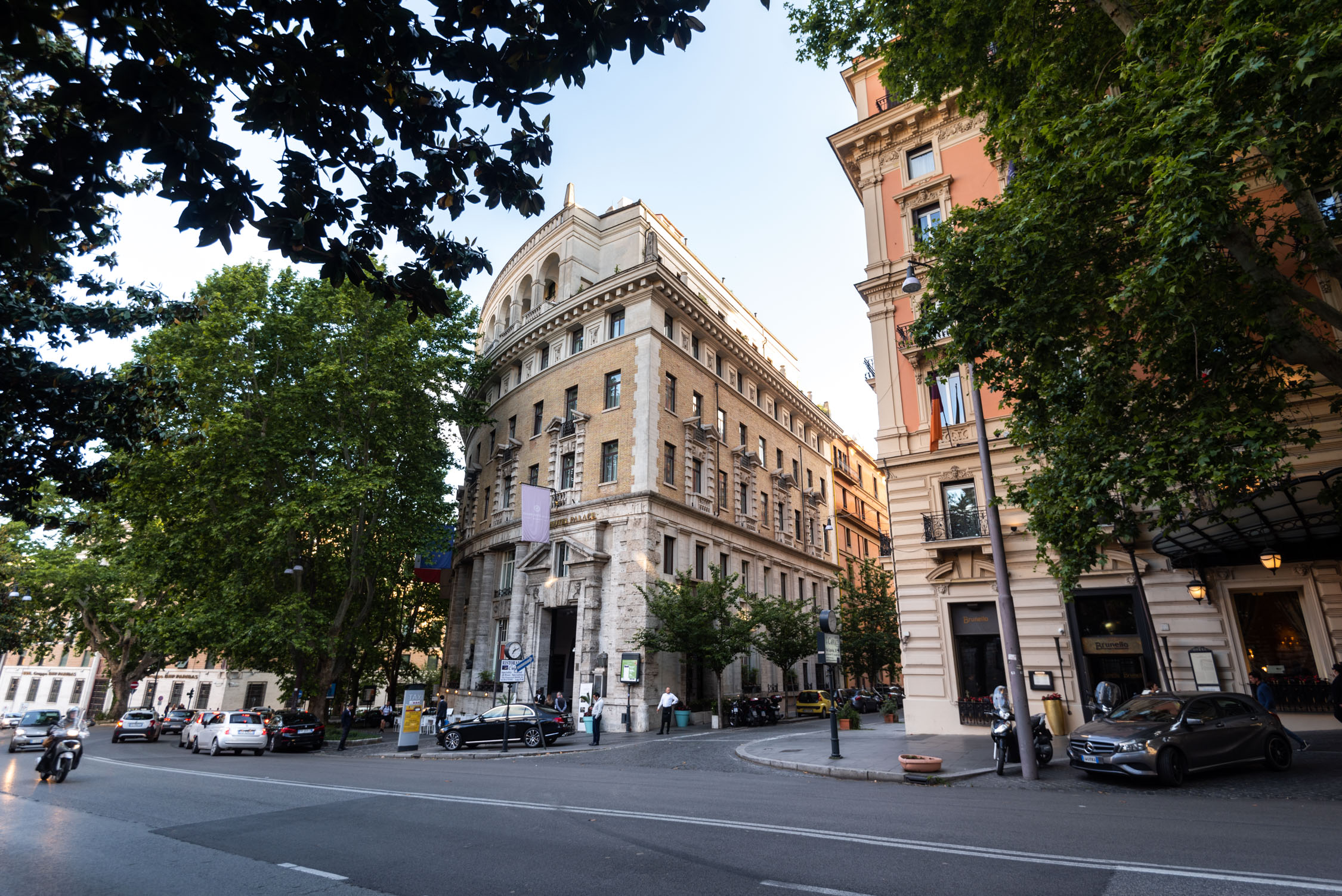 A wide street view of ornate European buildings with a large domed structure and parked cars under green trees.
