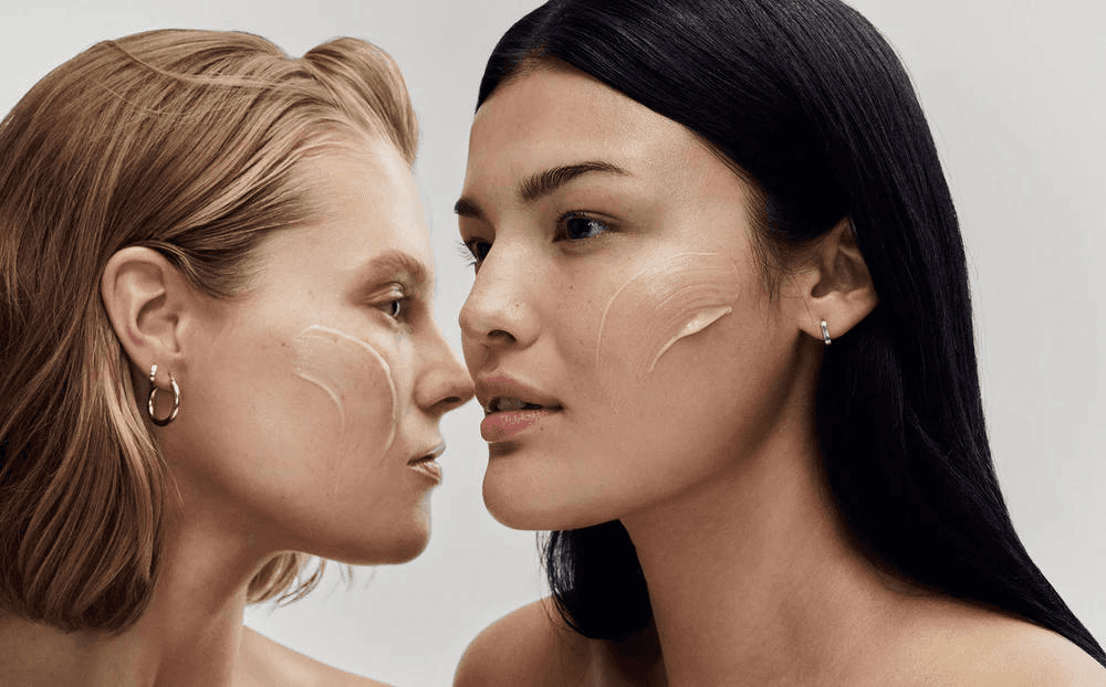 Two women standing closely together outdoors under a clear blue sky, looking directly at the camera, with blurred green leaves partially covering the foreground.