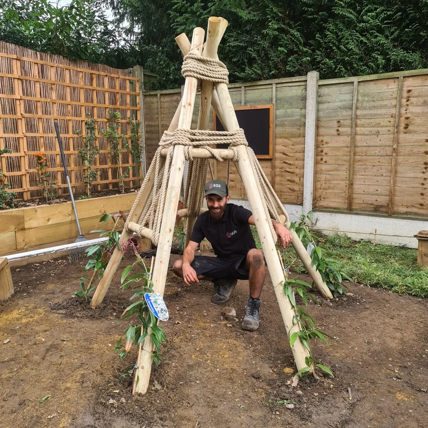 A child plays inside a wooden tepee structure, surrounded by a fenced garden area.