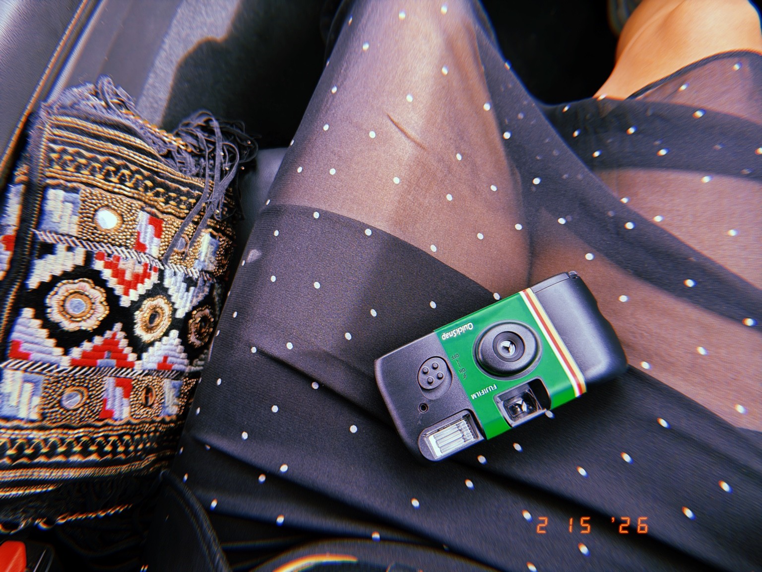 A green disposable camera on sheer dotted fabric beside an embroidered bag Photograph by Pranjali Shah.
