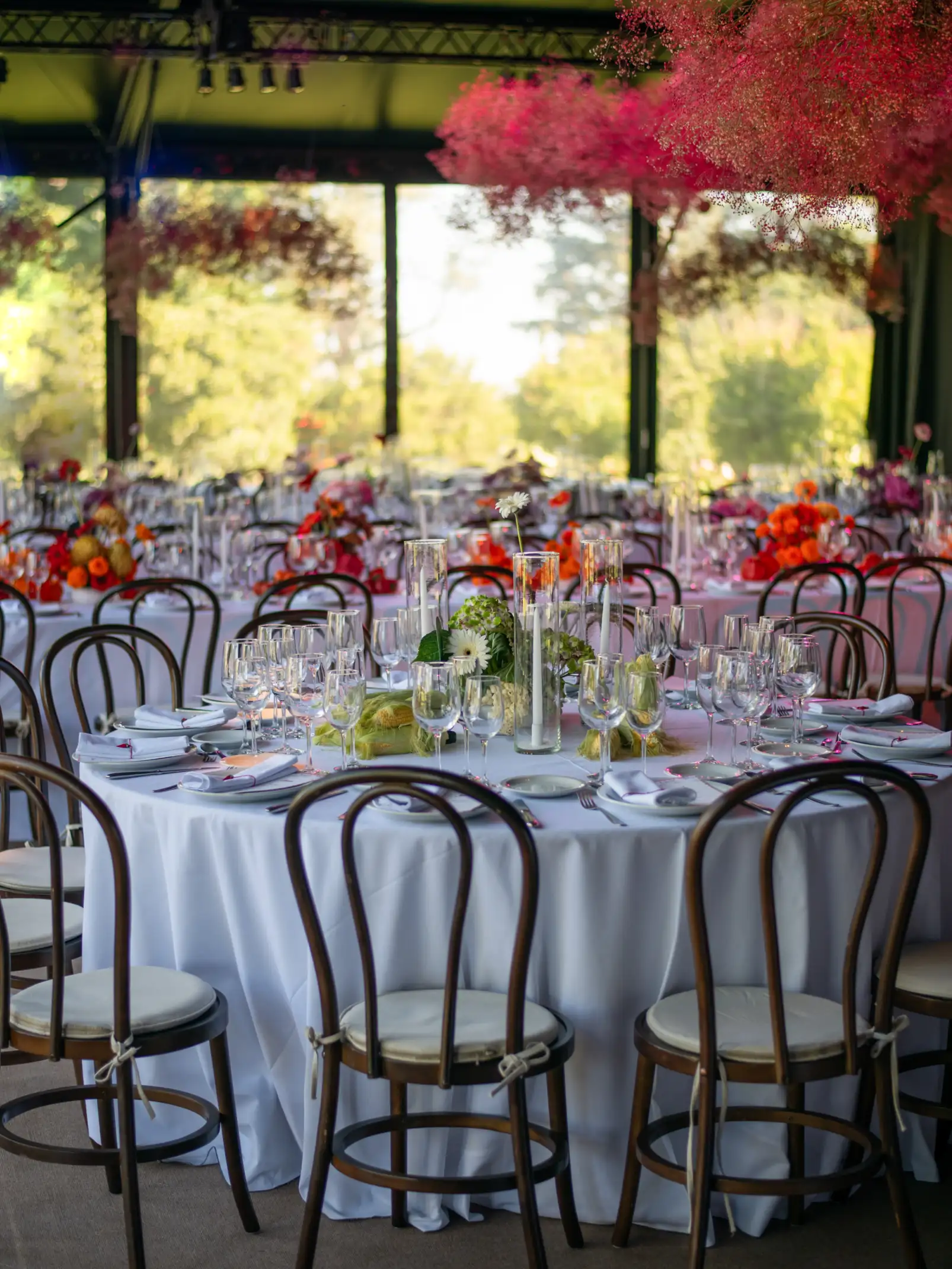 Salón con mesas redondas decoradas con flores y velas, bajo techo con instalación floral suspendida en tonos fucsia y coral.