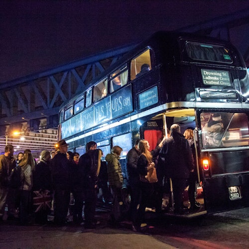 People queue to board a double-decker bus at night, with a sign for a tour company in the background and a bridge overhead.