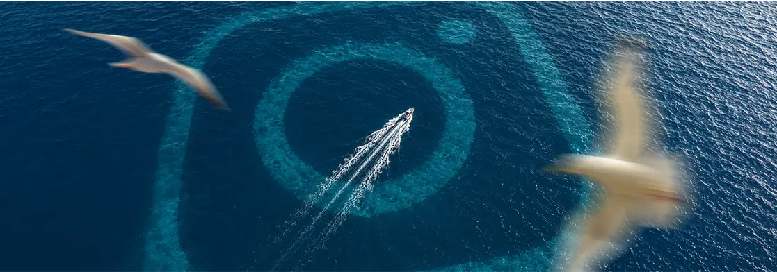 Vista aérea do oceano azul com o contorno gigante do logotipo do Instagram visível sob a água. Uma lancha branca cruza a parte inferior do logotipo deixando um rastro de espuma na água, enquanto duas gaivotas voam desfocadas em primeiro plano, uma de cada lado da imagem