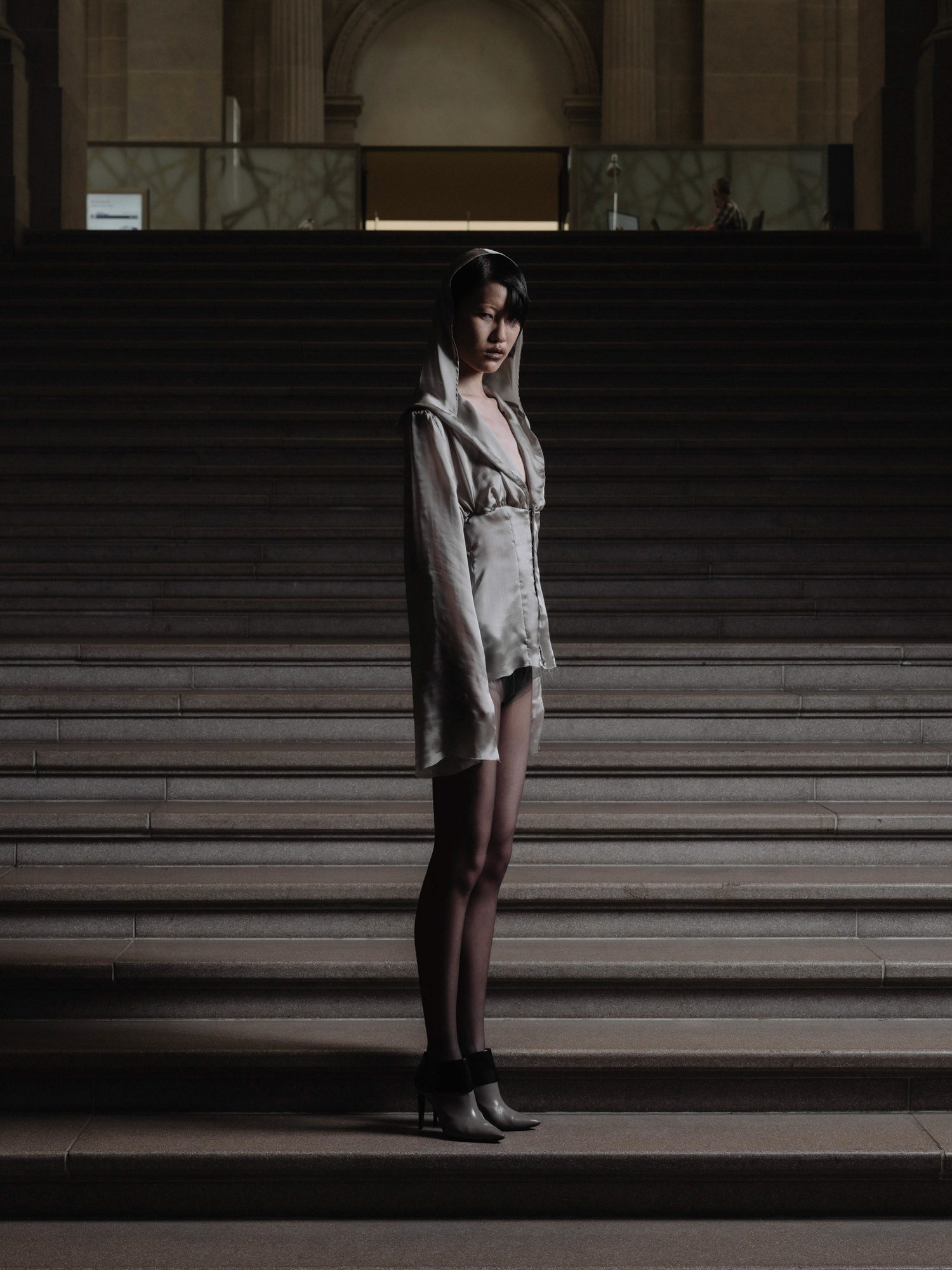 A person dressed in a stylish, silvery hooded outfit stands confidently on a staircase, blending gothic and avant-garde fashion elements amidst a dramatic indoor setting.
