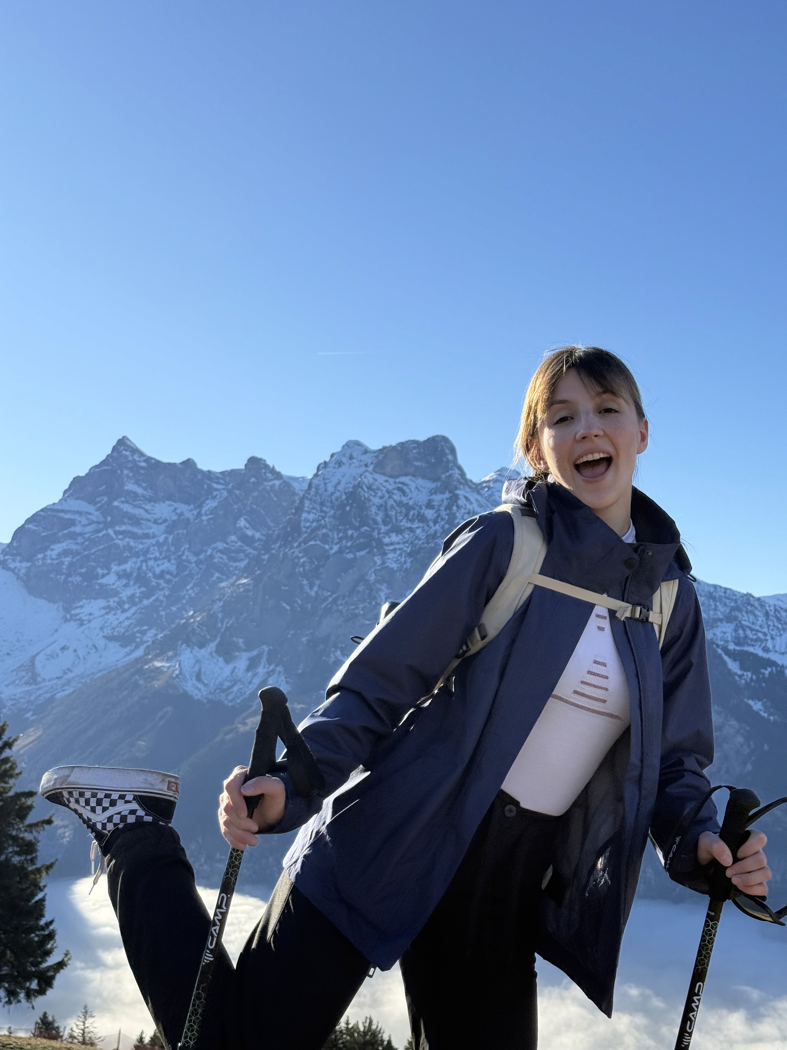 Una persona con una camiseta negra y pantalones de color claro está de pie en la cima rocosa de una montaña al atardecer, sosteniendo una cámara frente a su cara para tomar una foto. Un mar de nubes se extiende detrás de ellos, iluminado por la luz dorada del sol poniente.