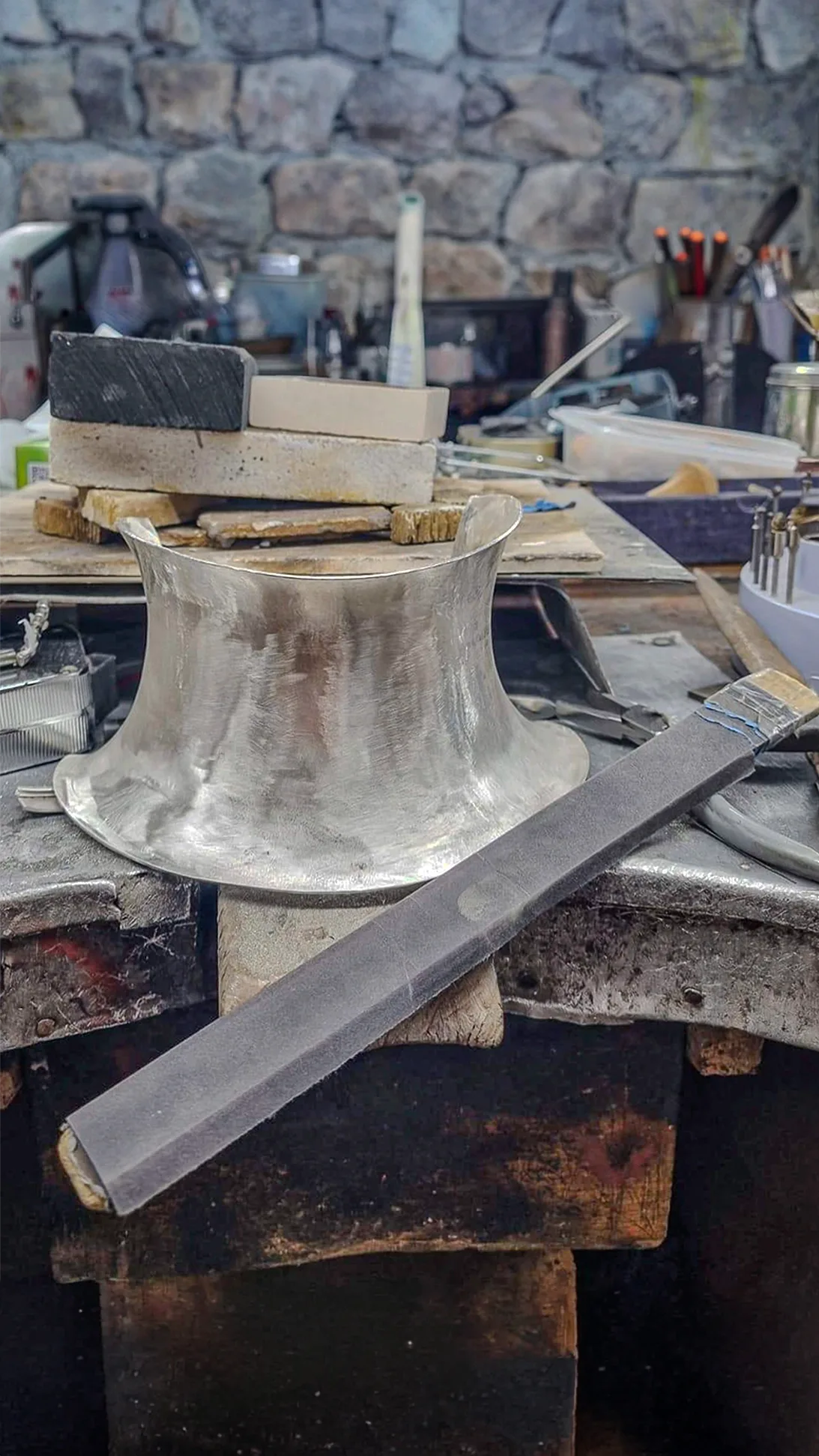 A high silver collar piece mid-craft sitting on a jeweler's workshop bench surrounded by artisan tools, files, and craft blocks, labeled "03 Form."