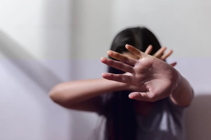 A imagem mostra uma pessoa com as mãos levantadas na frente do rosto, em um gesto de proteção ou defesa. A pessoa está desfocada, com o foco principal nas mãos que parecem estar bloqueando algo, sugerindo uma tentativa de se proteger de uma ameaça. A cena transmite uma sensação de vulnerabilidade e resistência, e o ambiente ao redor parece ser simples e neutro, com pouca iluminação, o que contribui para o tom sombrio e emocional da imagem.