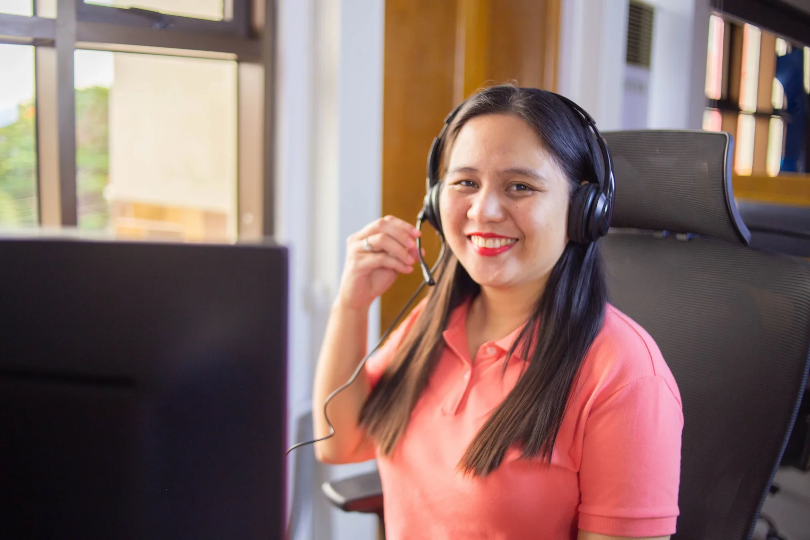 A woman wearing a headset smiles while sitting at her desk in a customer support or call center environment.