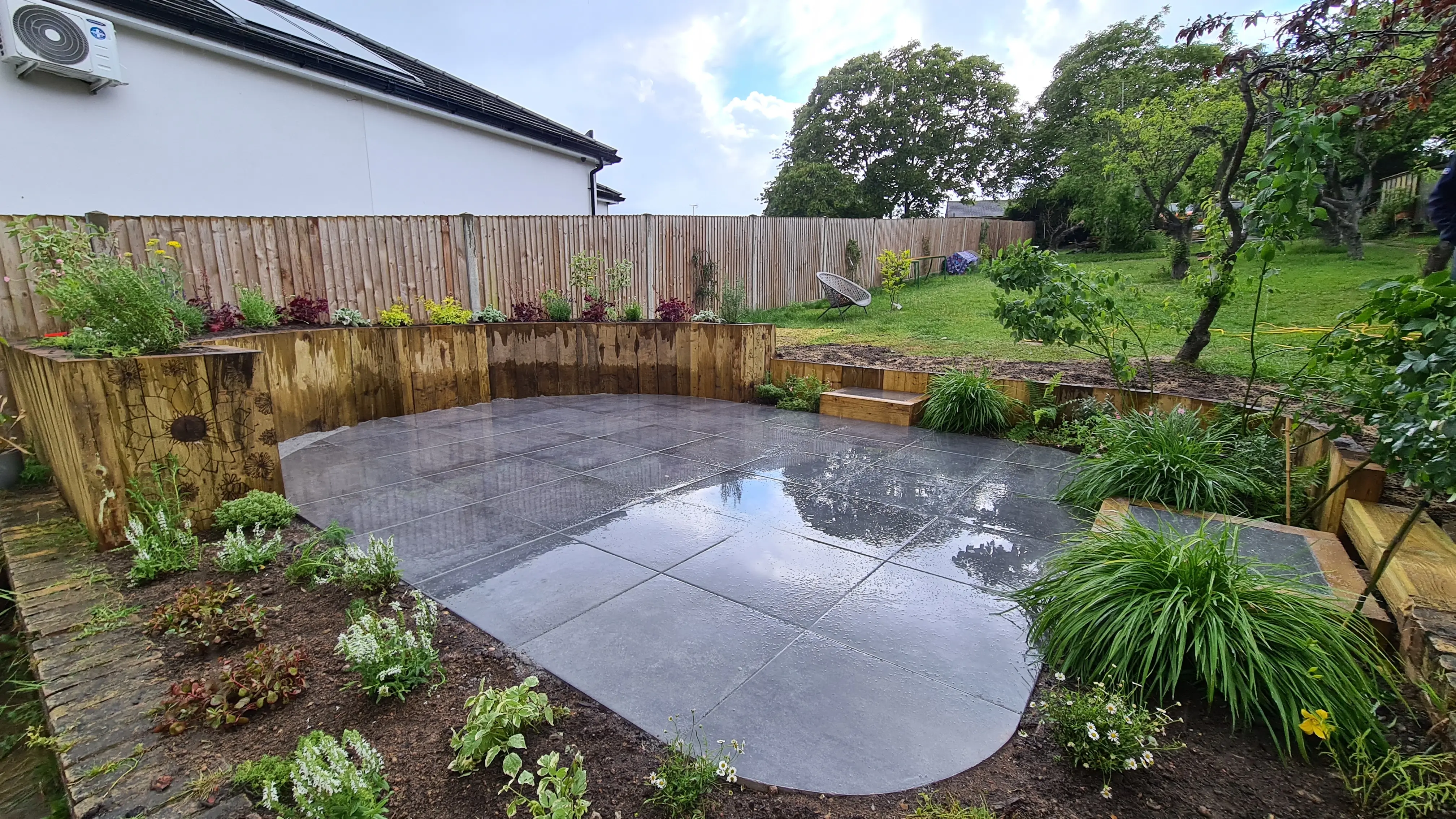 A serene garden with a small pond, surrounded by greenery and a wooden fence, under a partly cloudy sky.