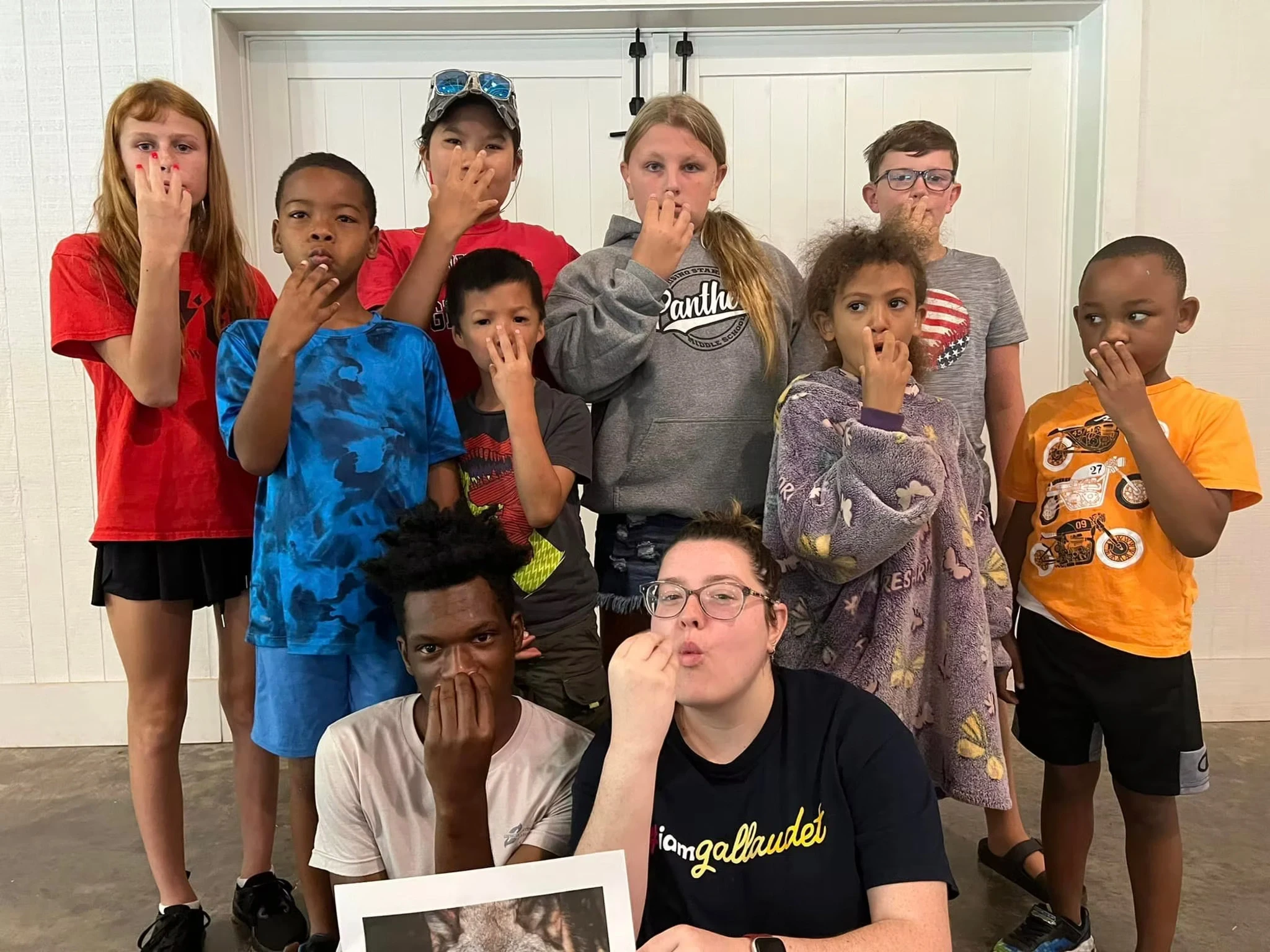 A group of campers and a staff are posing for a photo. They are all signing WOLF as the staff member holds a picture of a wolf.