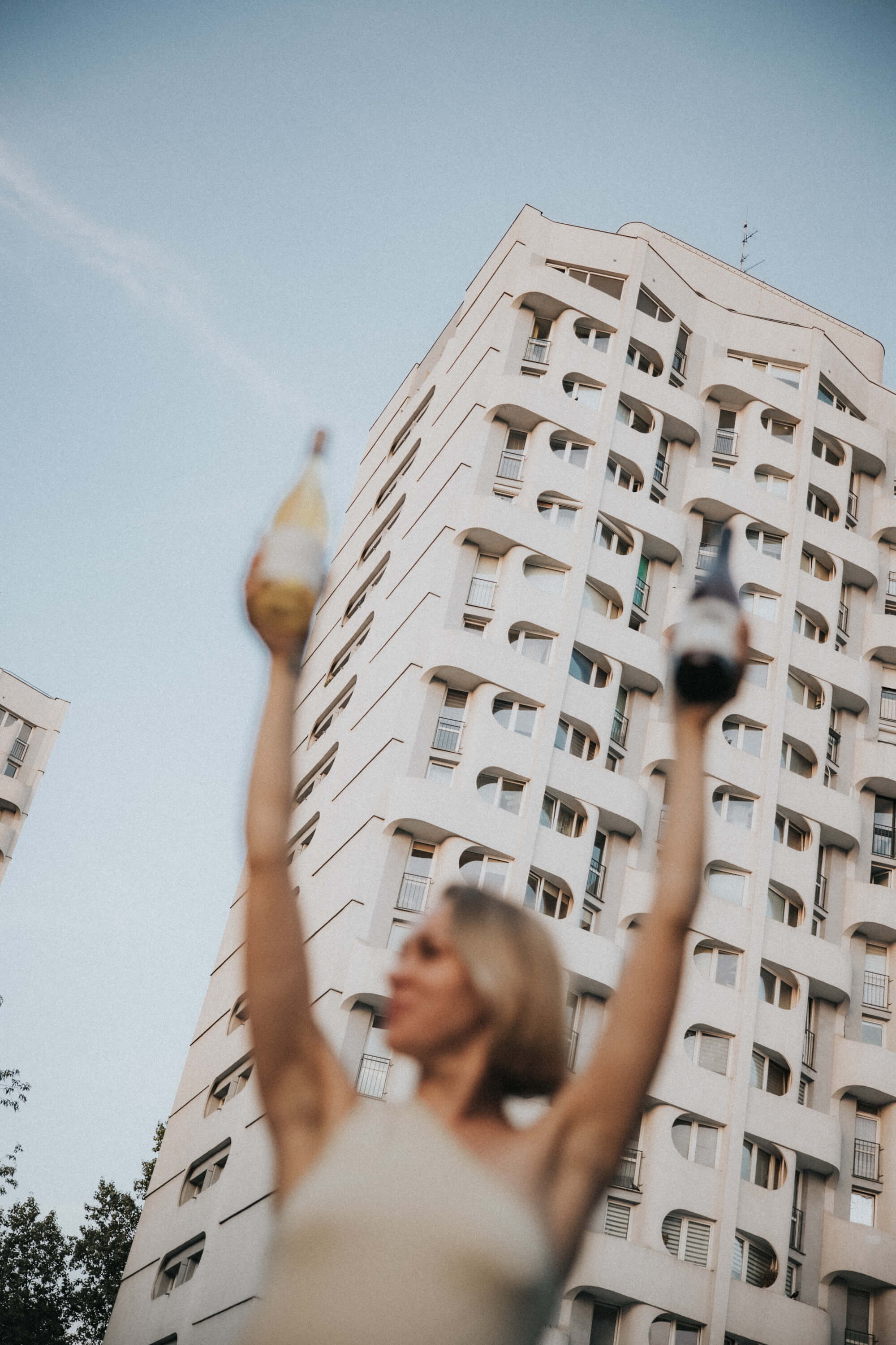 A woman stands outdoors, raising her arms while wearing gloves, with a tall building in the background.