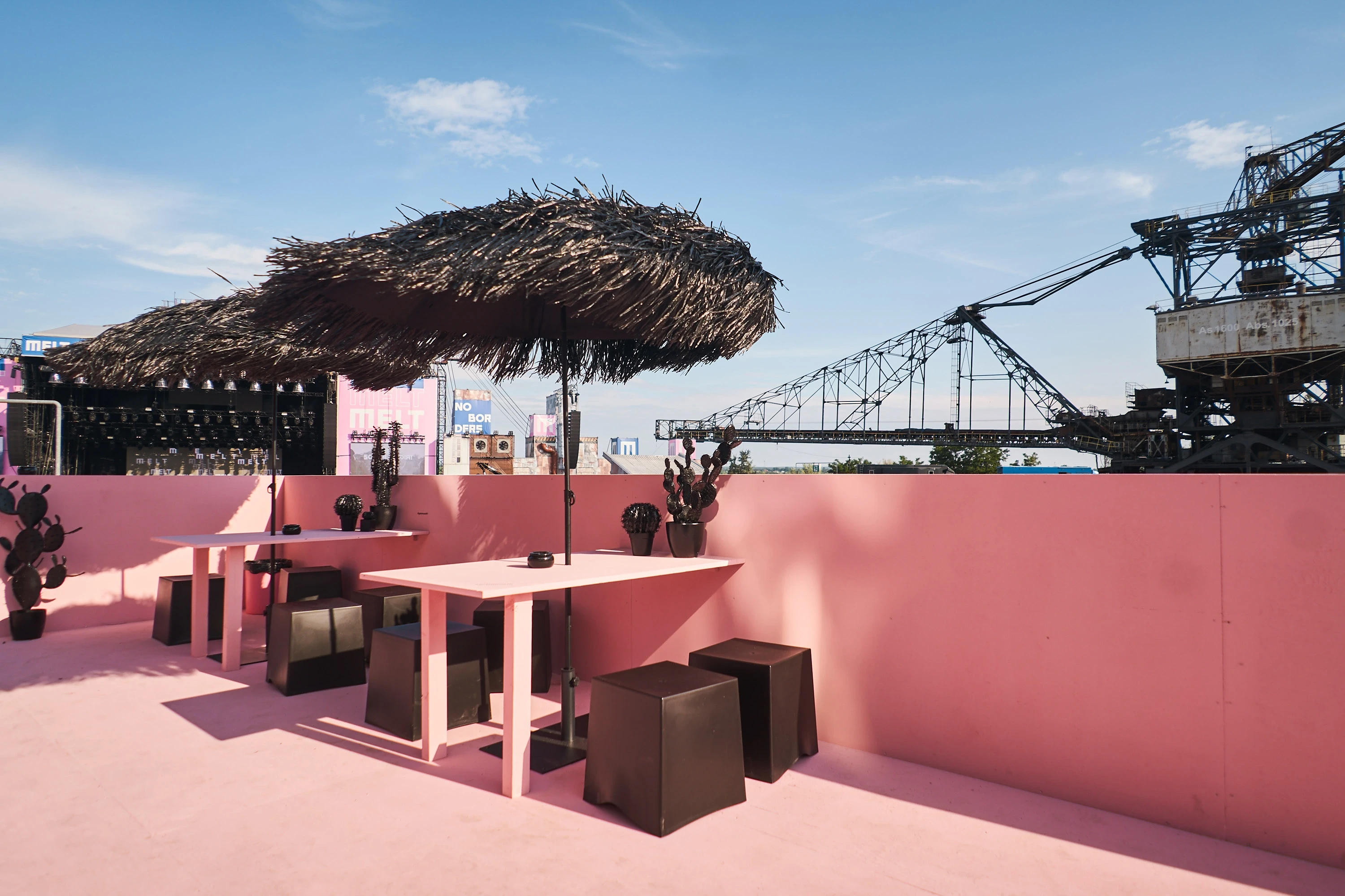 Pink roof terrace of the Klarna platform with palm-frond parasols, high tables and black stools overlooking the festival grounds.