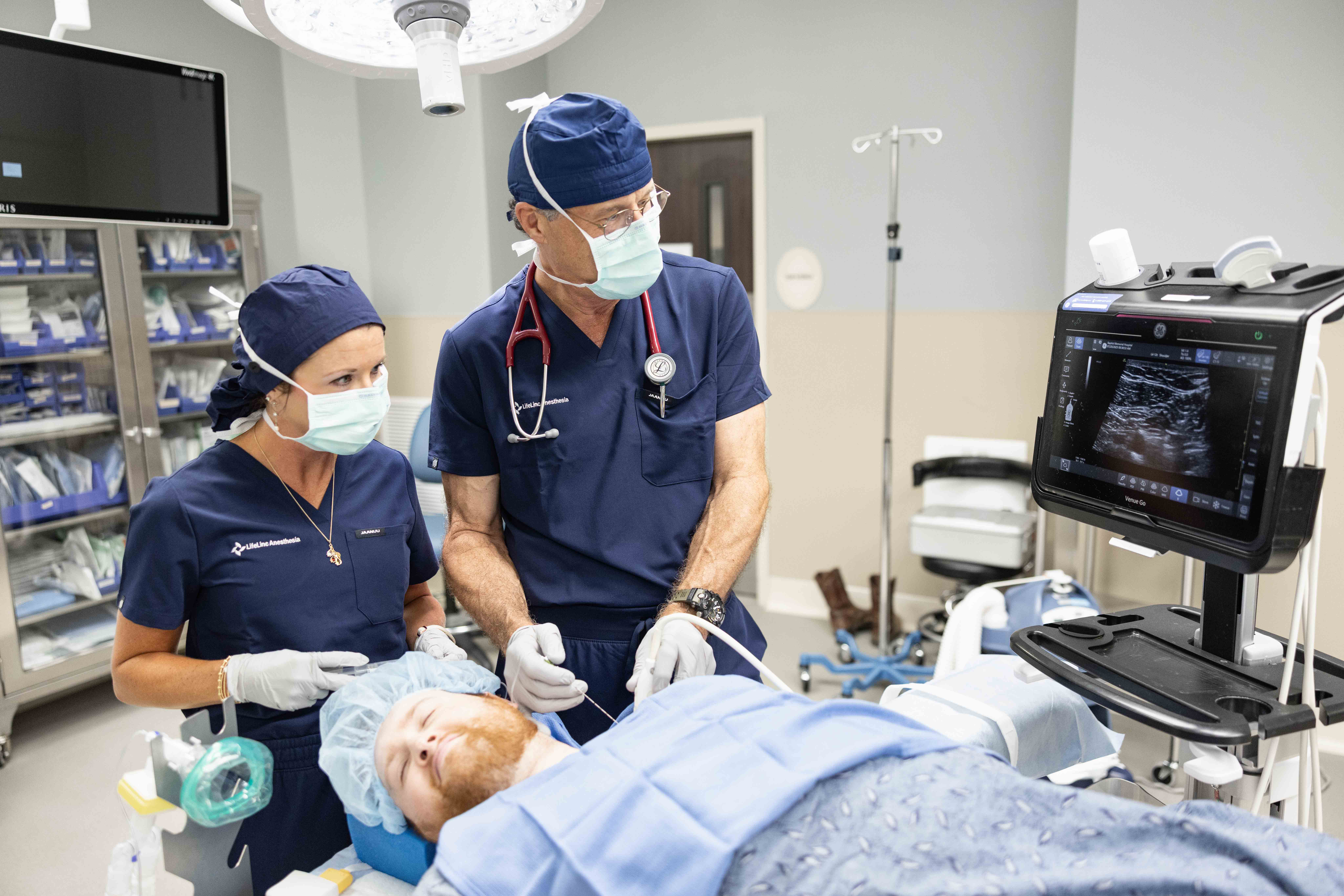 Two anesthesia providers in scrubs and surgical masks prepare a patient for a procedure in a hospital operating room