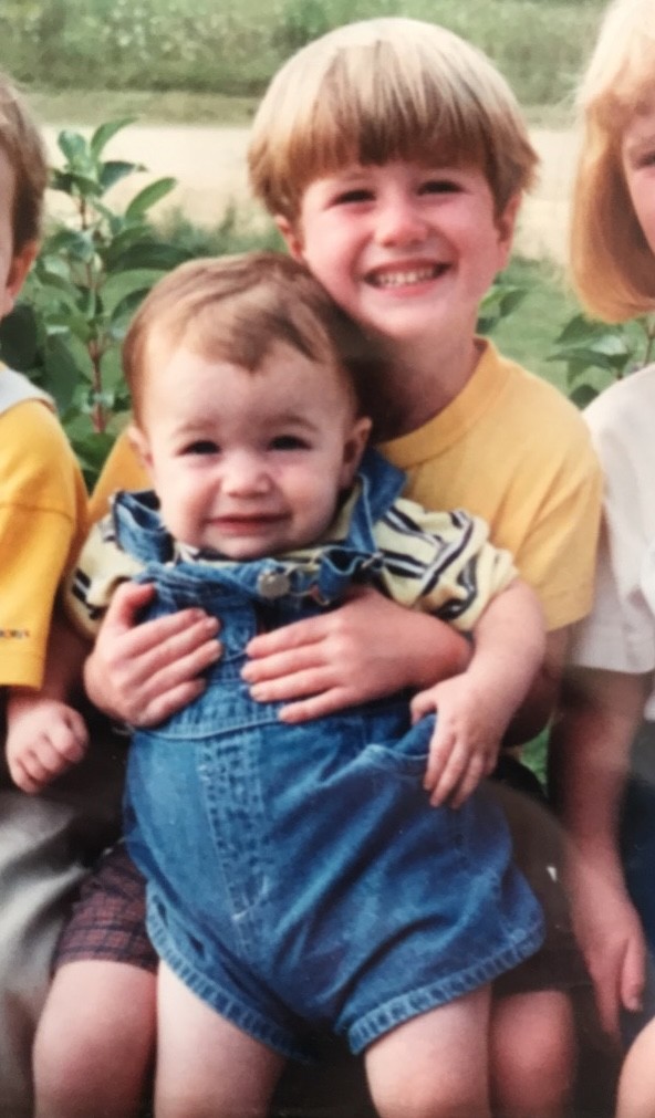 A smiling man with curly hair is sitting at a desk, looking at the camera, with a laptop and a lamp in the background.