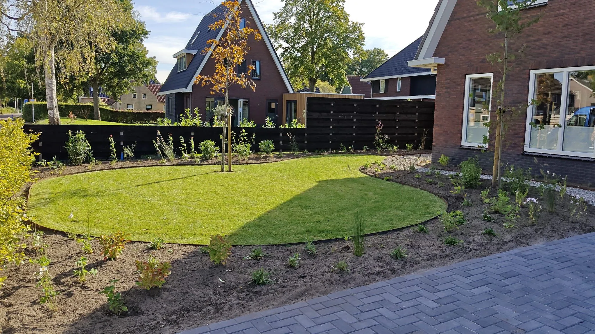 Nieuw aangelegde kindvriendelijke tuin met strak gazon, jonge bomen en planten, omgeven door een zwarte houten schutting bij een nieuwbouwwoning.
