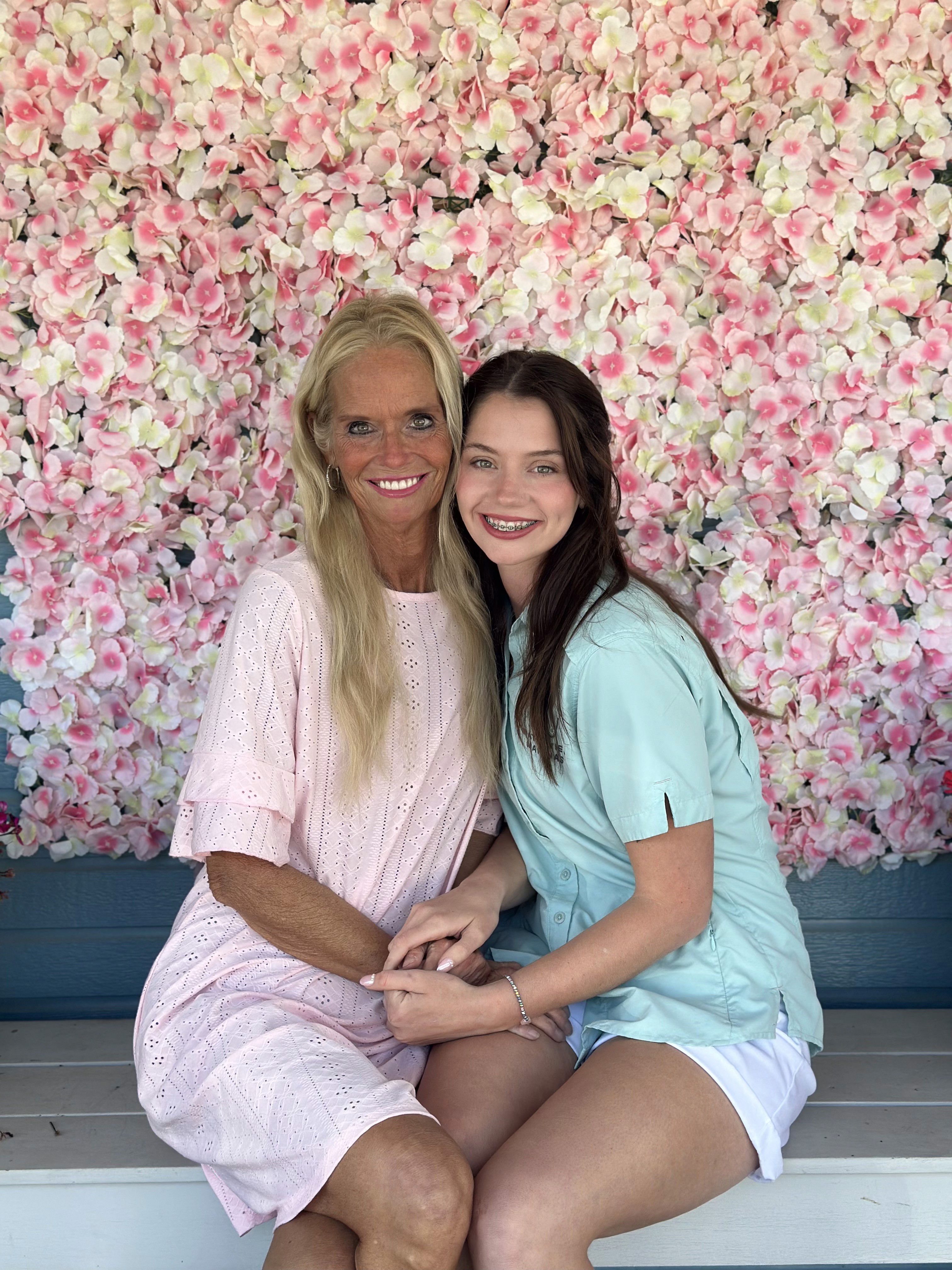 Two people sit closely on a white bench against a vibrant backdrop of pink and white flowers, smiling warmly at the camera, creating a cheerful and inviting scene.