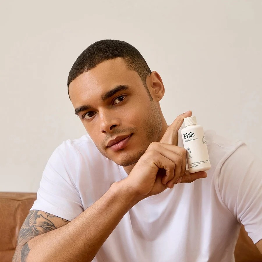 A model holding a 100ml travel size VIVOMER bottle of Phil's daily facial cleanser. The compact format proves the material's versatility for portable shelf-stable skincare liquids.