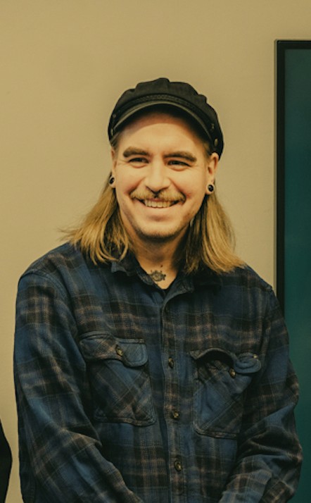 John smiling and dressed in blue shirt and a black hat. Photo by Tero Marin.