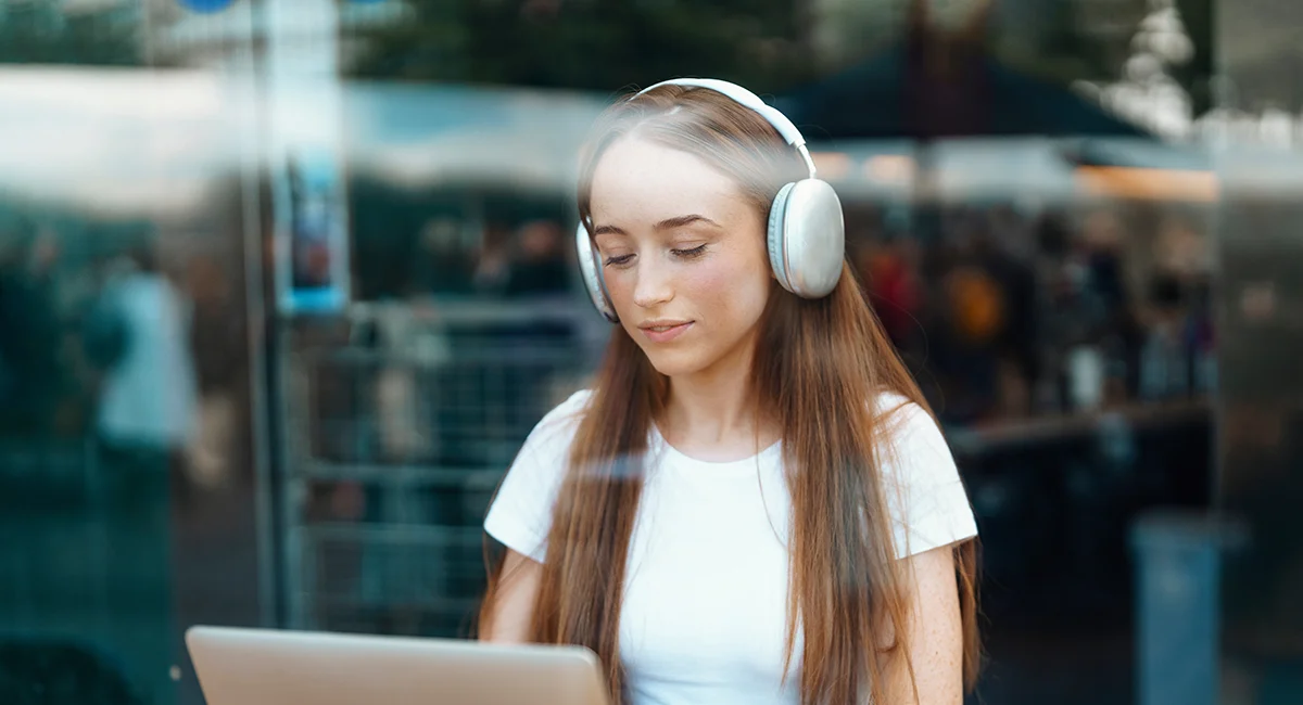 A woman with long hair, wearing headphones, focused on her laptop outdoors.