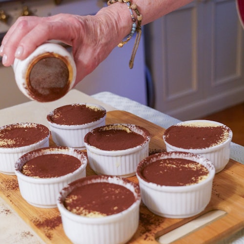 A hand sprinkling cocoa powder over eight ramekins filled with dessert, placed on a wooden board in a kitchen.