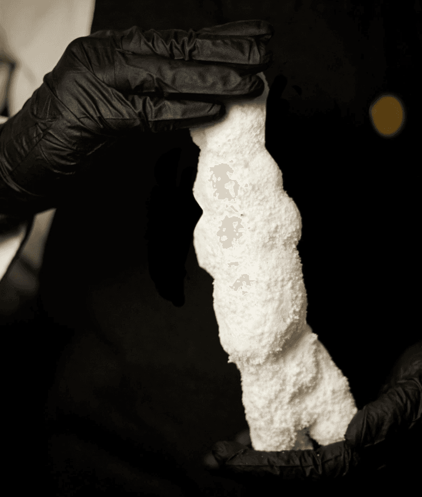 A chef wearing black gloves holds a tall, sculpted piece of white salt, shaped like a snowman, against a dark background.