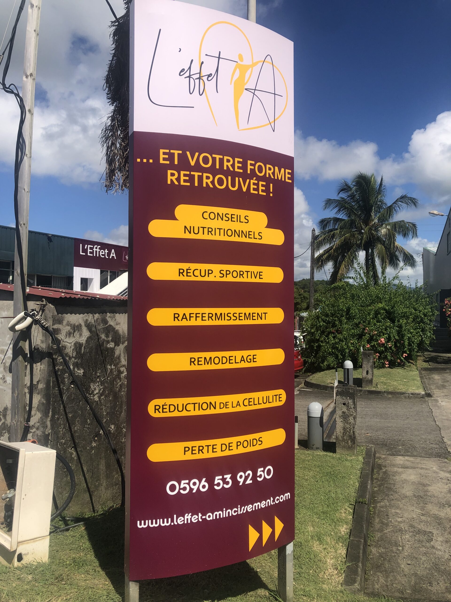 Grand panneau publicitaire pour « L'Effet A » sur fond de ciel bleu, proposant des services tels que des conseils nutritionnels et des programmes de perte de poids. Palmiers et végétation à proximité.