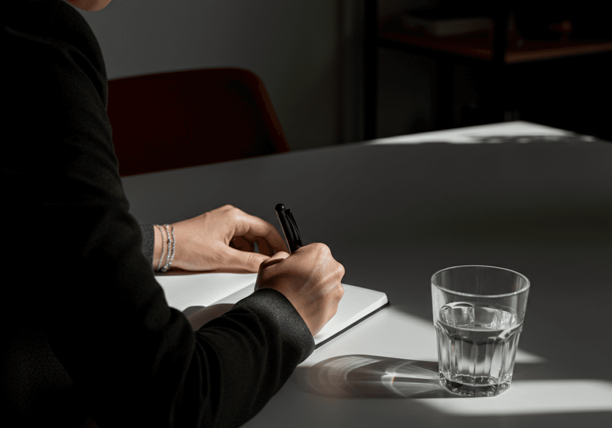 Person writing in a notebook at a table with a glass of water, creating a focused and calm atmosphere.