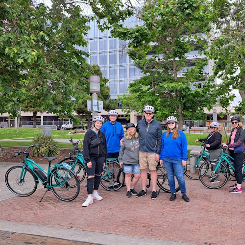 一群戴着头盔的人站在自行车旁，背景是建筑物和树木。