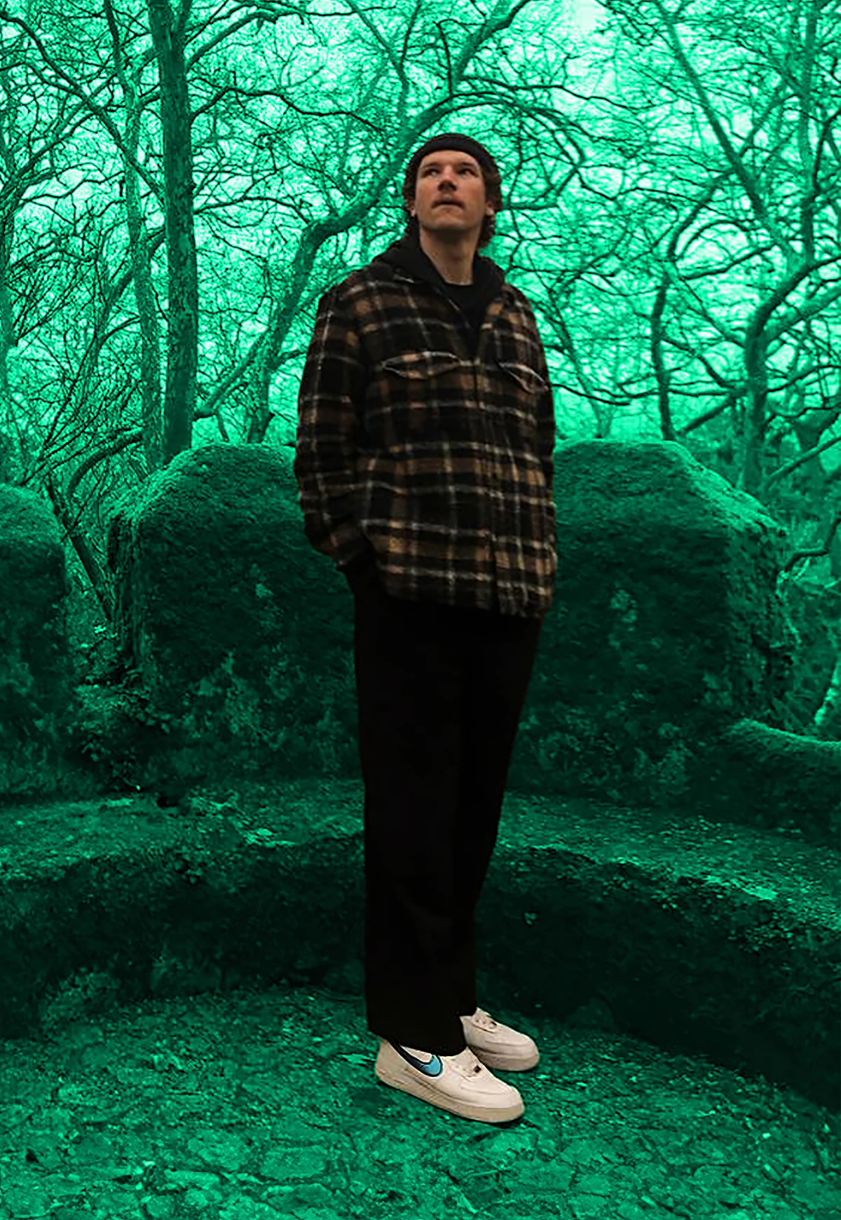 Ein Mann steht in einer surreal wirkenden, grün getönten Waldlandschaft mit Moos bedeckten Steinen und Bäumen. Er trägt eine Mütze, ein kariertes Hemd, schwarze Hosen und helle Sneaker. Sein Blick ist nach oben gerichtet.