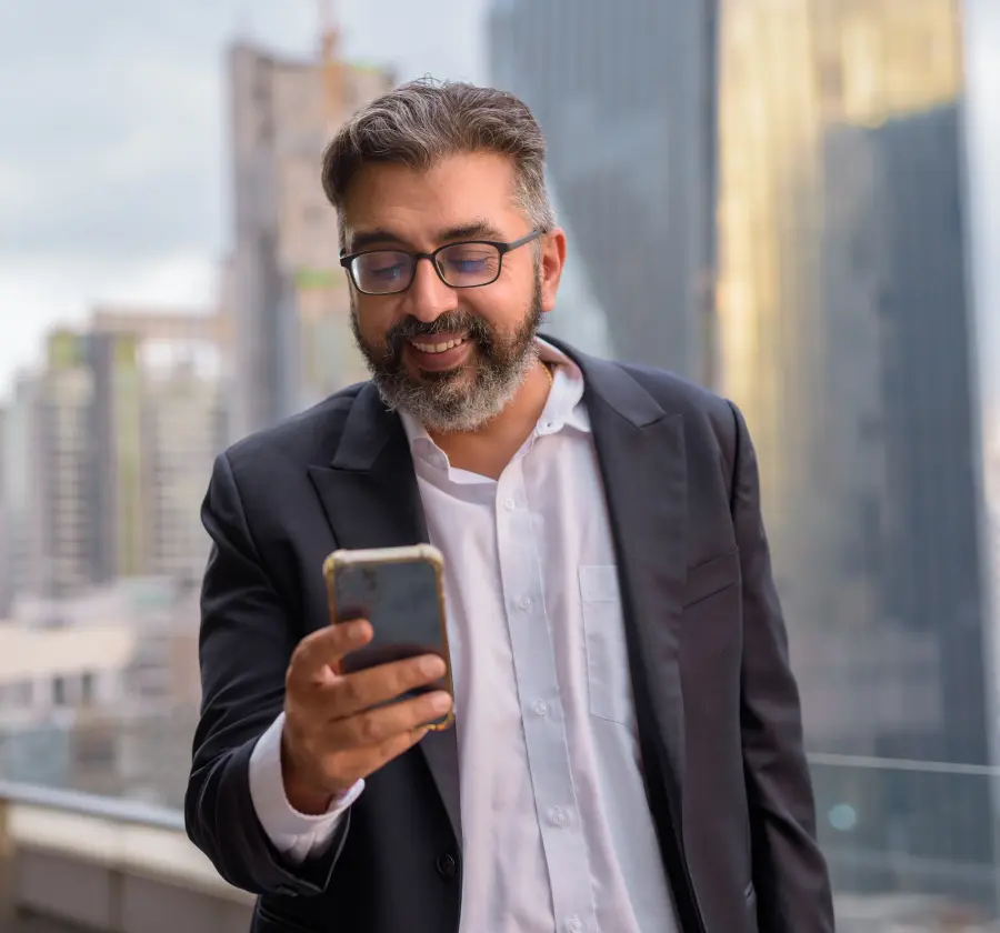 A middle-aged Indian businessman in a suit smiling while using his smartphone outdoors, representing the professional and convenient use of digital health platforms for managing chronic conditions.