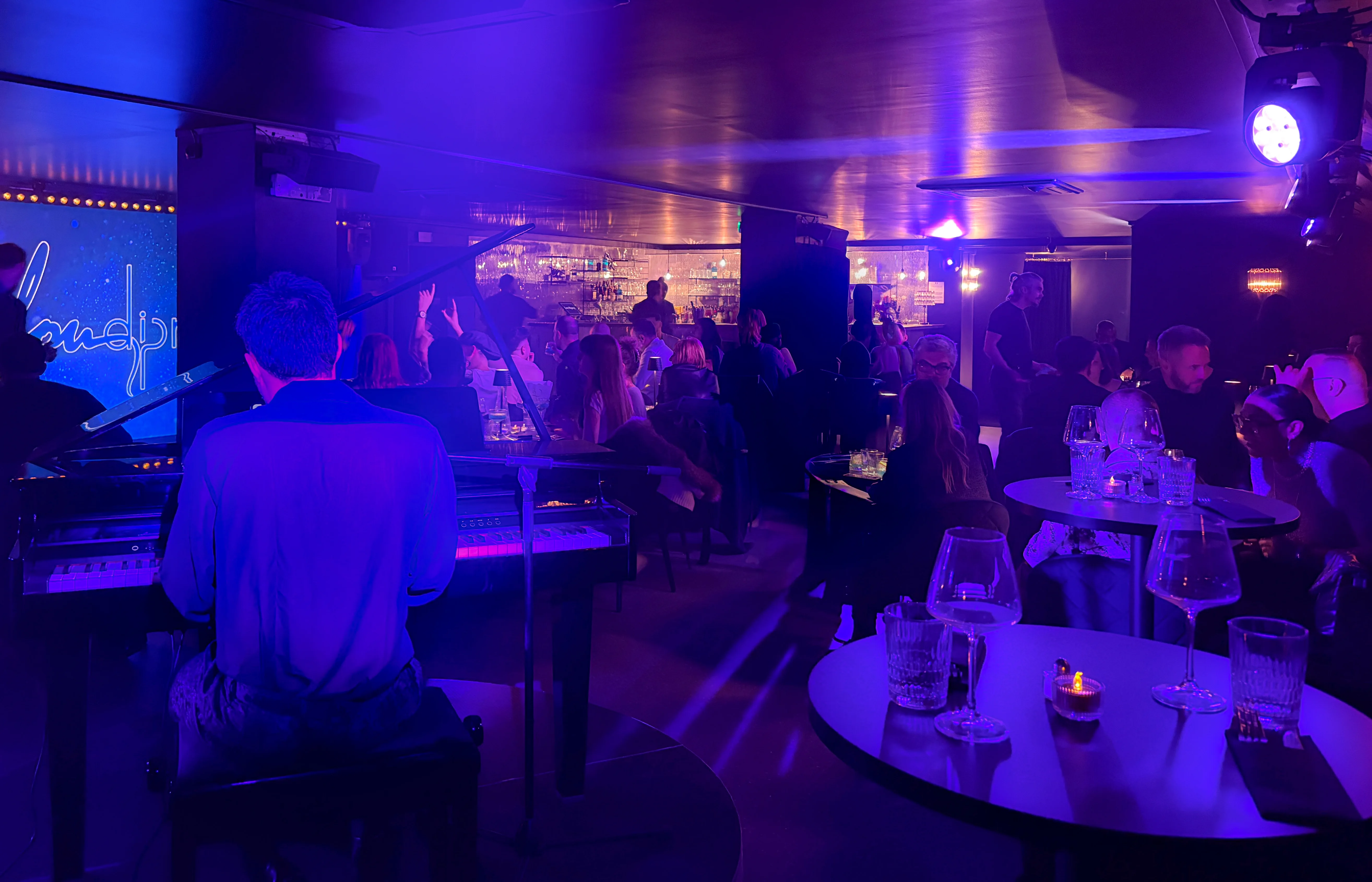 Atmospheric wide shot of the Lou Diprey cabaret interior, bathed in deep purple and blue stage lighting. Blurred patrons are seated at tables in the foreground, facing a stage area with focused spotlights and a prominent grand piano