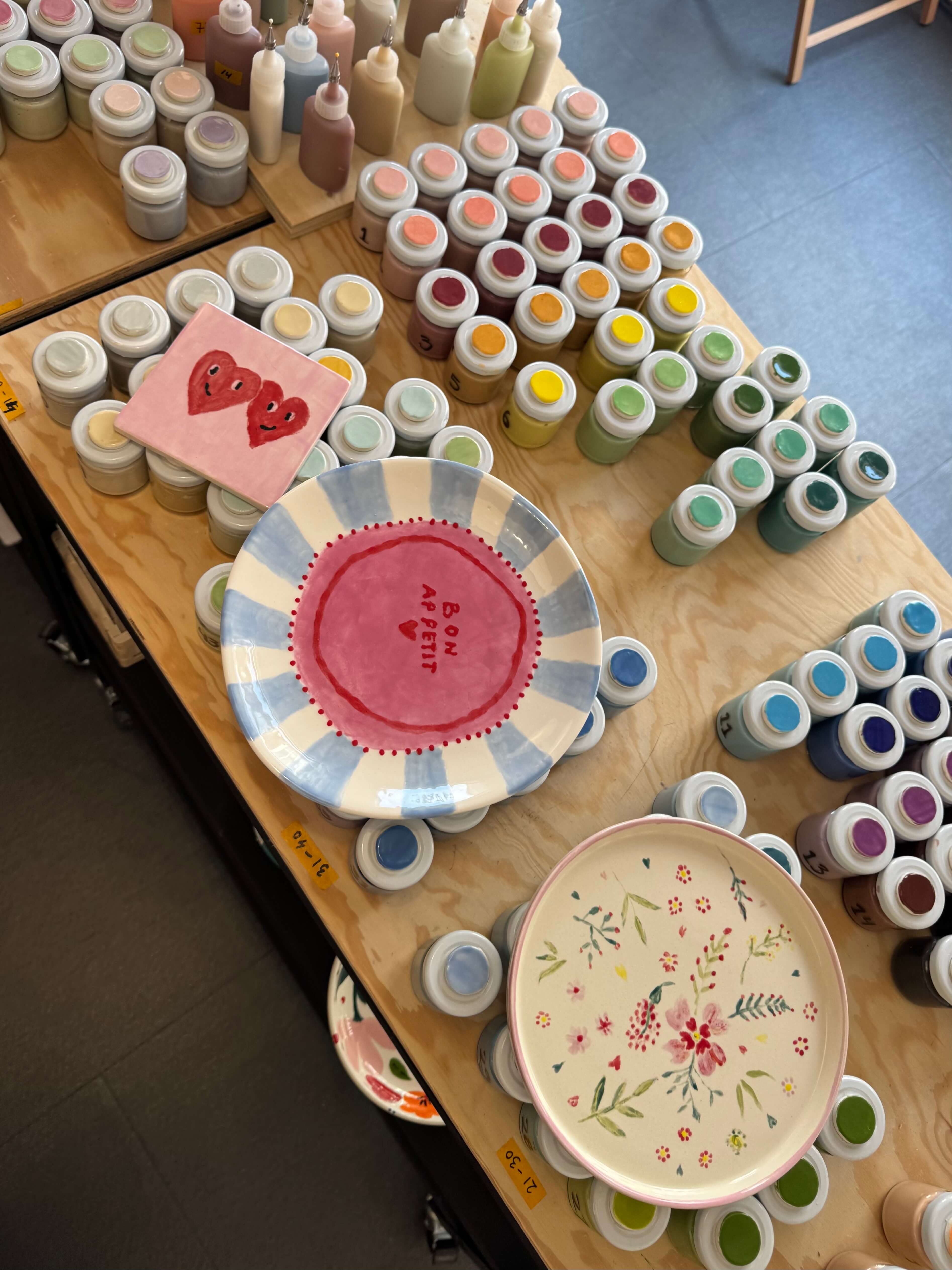 A table full of underglaze paints with two painted plates and one tile 