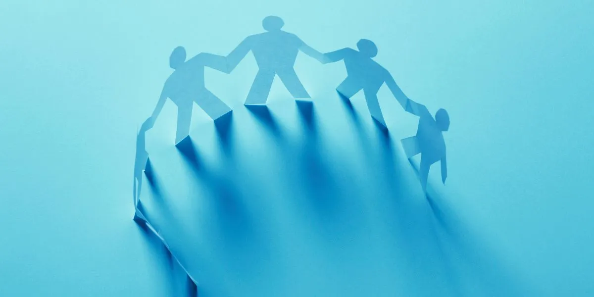 A man stands while two people sit at a table in an office setting, smiling and engaged in conversation. The mood is friendly and collaborative.