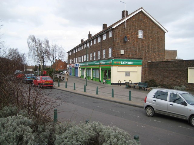A smiling man with curly hair is sitting at a desk, looking at the camera, with a laptop and a lamp in the background.