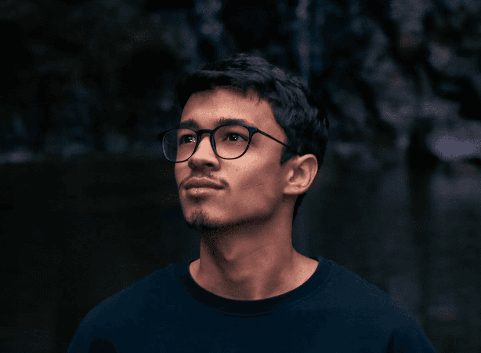 Un jeune homme portant des lunettes regardant vers le haut, avec un arrière-plan sombre et flou. L'éclairage est doux et tamisé. Developpeur web 974