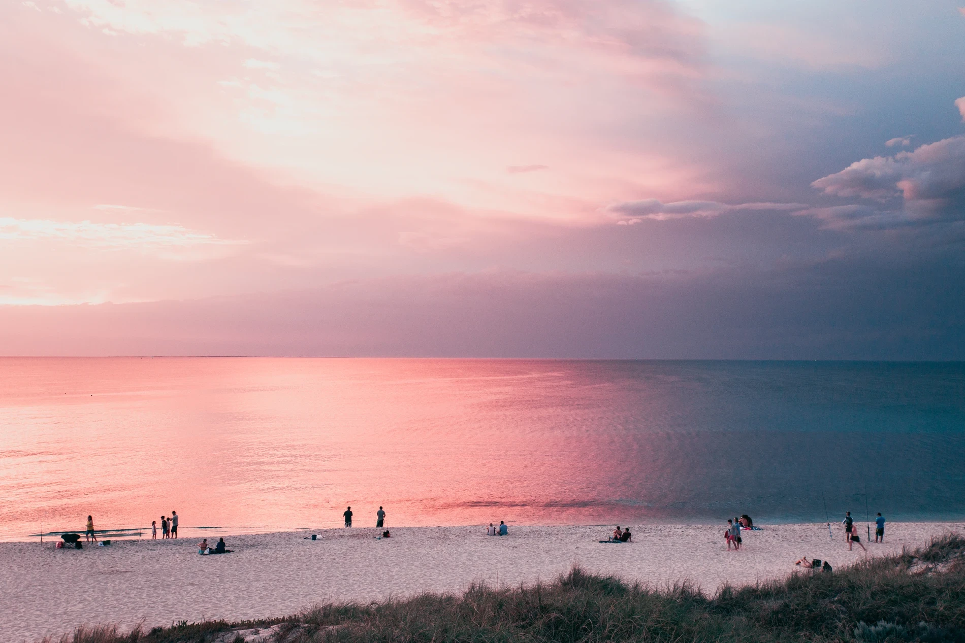 A pink and lilac sunset over a calm sea, figures dotted along the shoreline