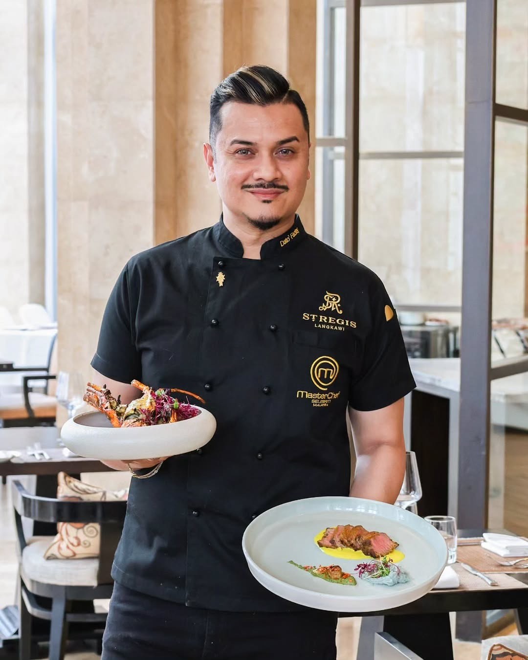 A chef in a black uniform holding two beautifully presented plates of food inside a restaurant setting at The St. Regis Langkawi.