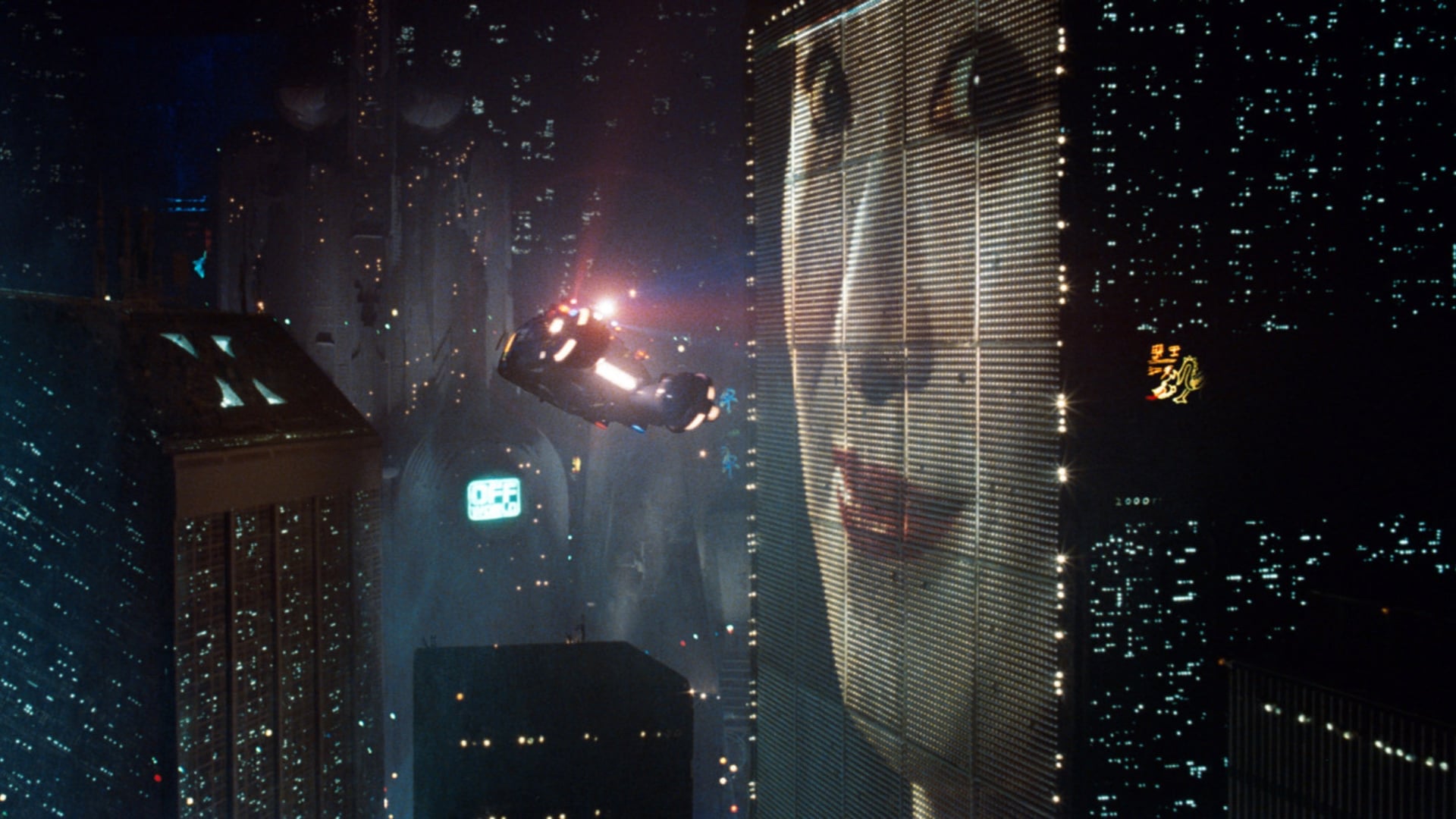 Scene from Blade Runner film. A futuristic cityscape at night featuring skyscrapers with bright lights and digital advertisements. A flying car hovers near a building with a large electronic billboard displaying a woman's face.