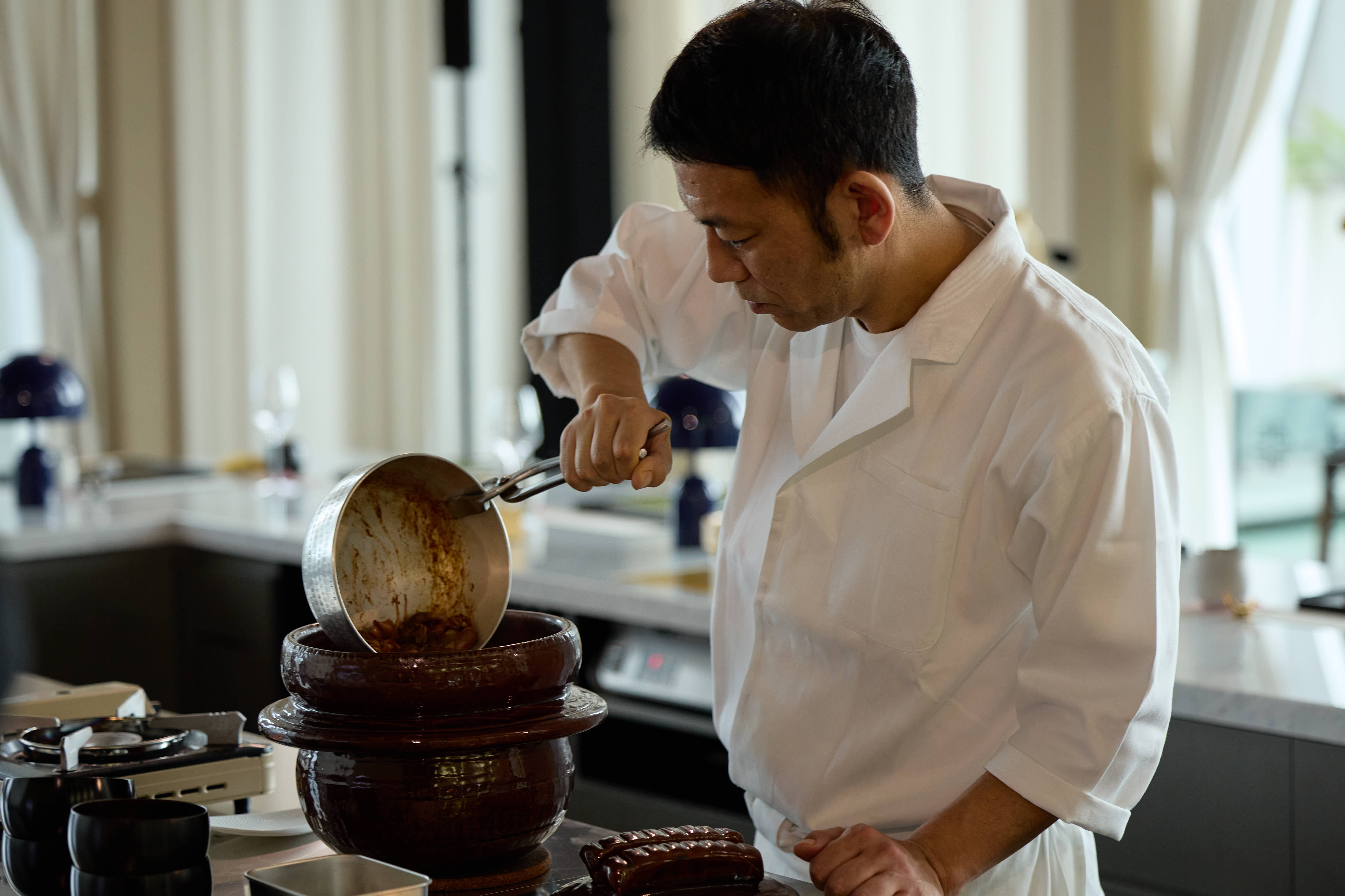 Michelin two-star Chef Yozo Ueda, dressed in a white chef’s coat, focused intently as he pours ingredients from a small pot into a large, traditional ceramic bowl during the Taipei residency.