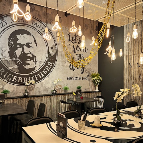 A restaurant interior with hanging pendant lights, rustic decor, wall art, and floral arrangements on tables and shelves.