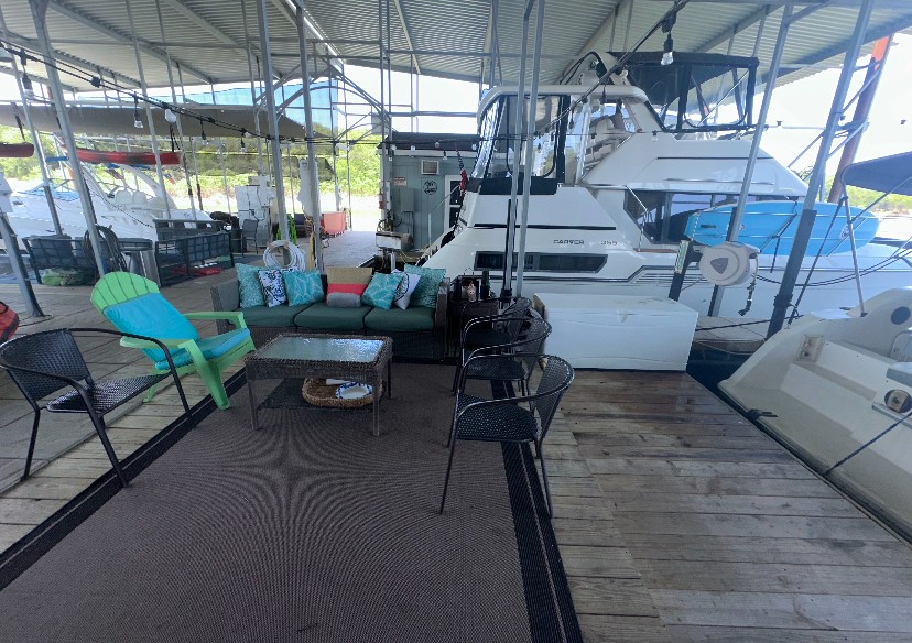 A cozy marina dock setup features a colorful lounge area with a teal chair, cushioned sofa, and a coffee table, surrounded by several elegant boats under a covered structure.