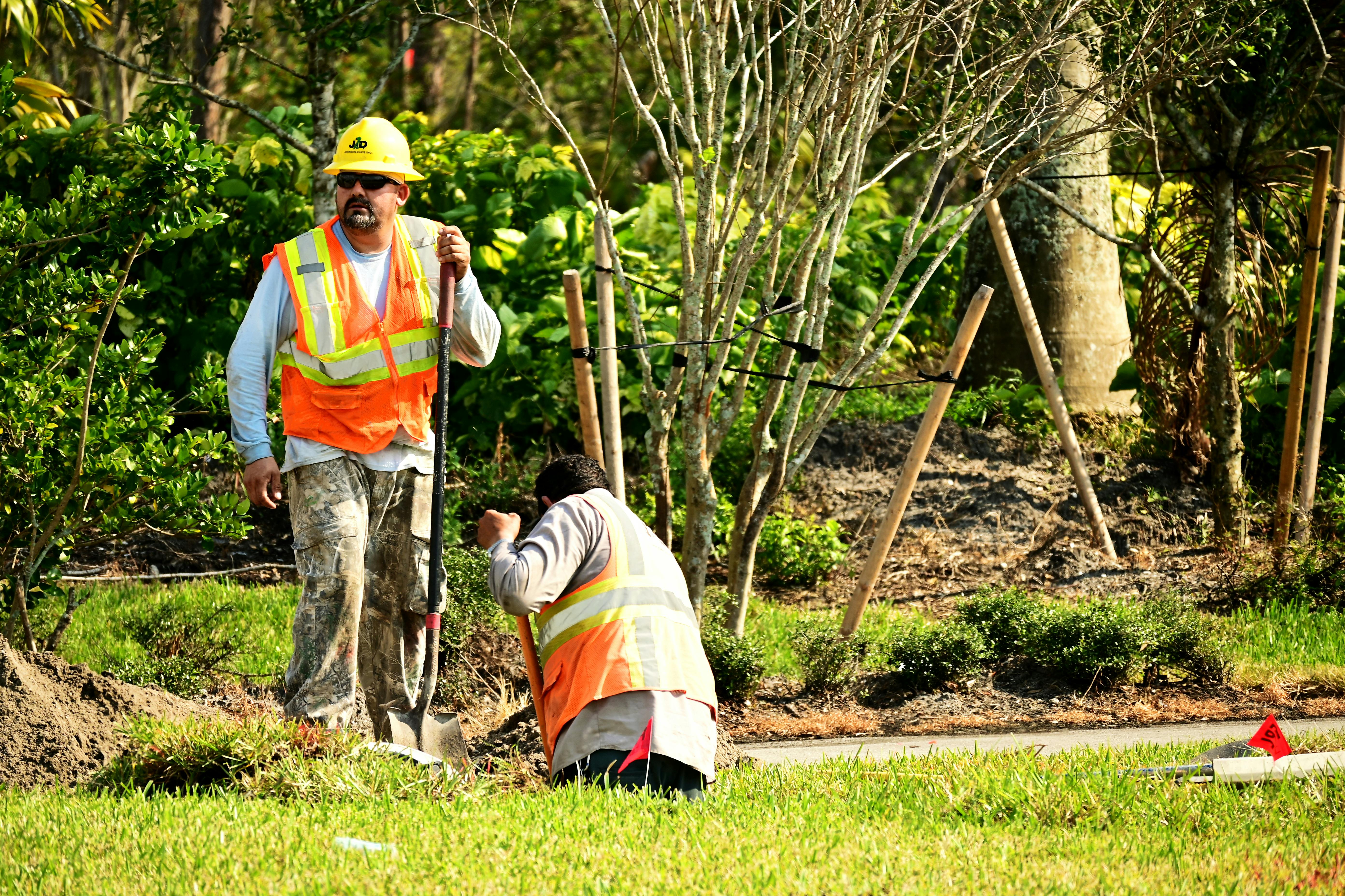 Worker in a high-visibility vest digging outdoors near trees.