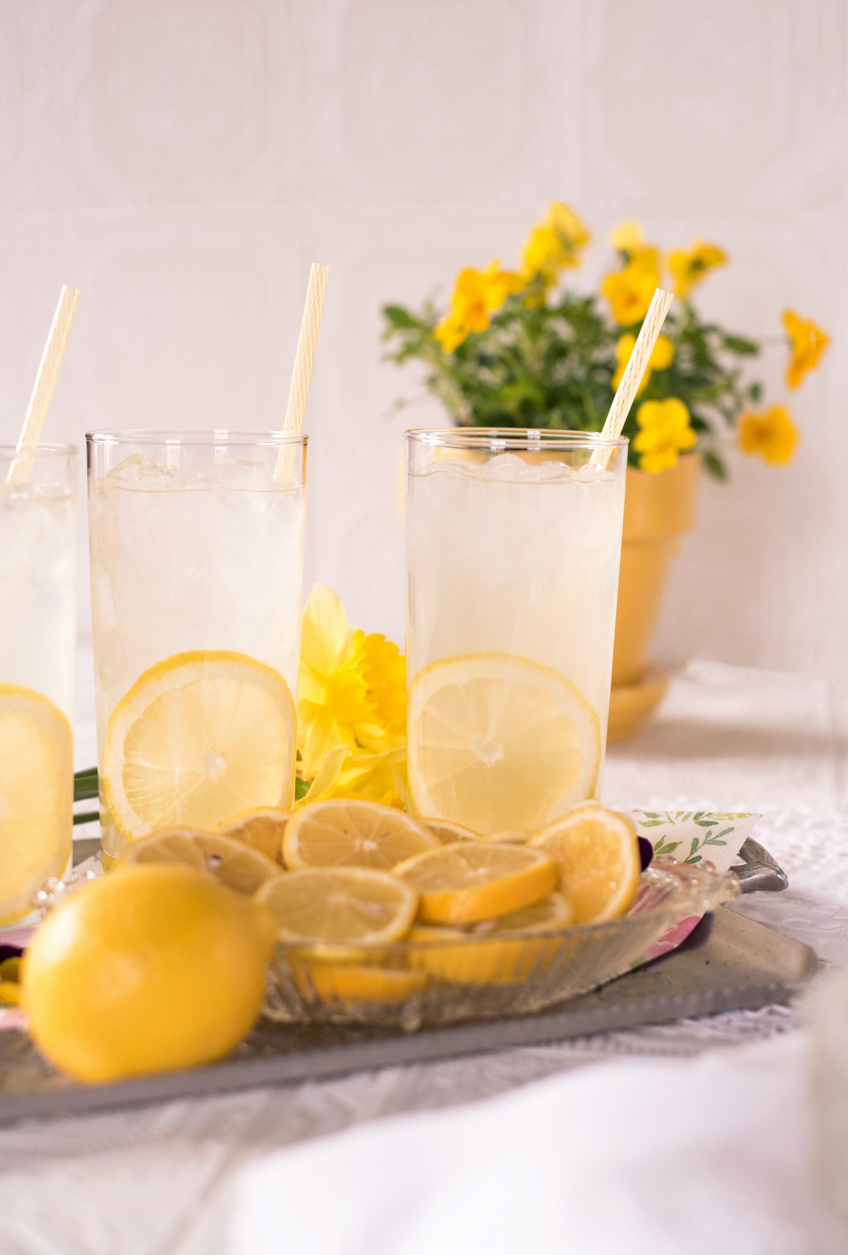 Lemonade surrounded by sliced and whole lemons on a metal tray with bright yellow daisies in the background.