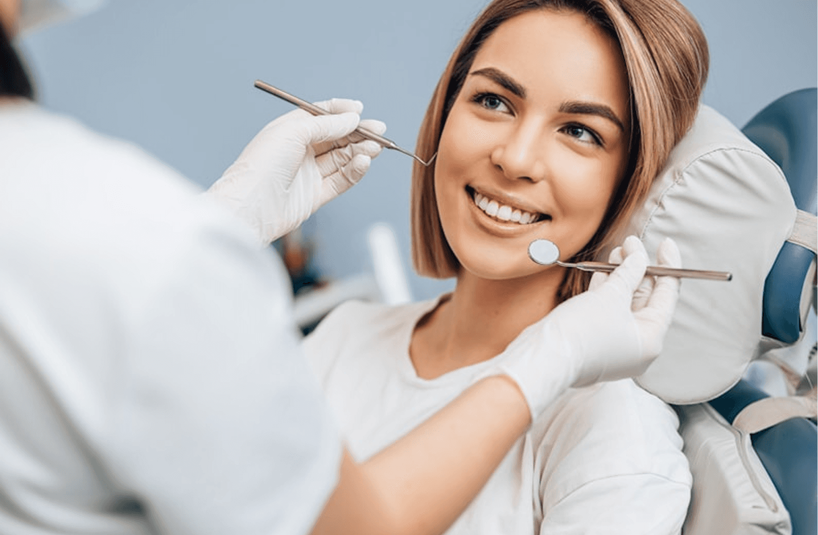 A woman sits in a dental chair, smiling as a dentist checks her teeth with a dental mirror and probe, highlighting the importance of dental care and oral health.