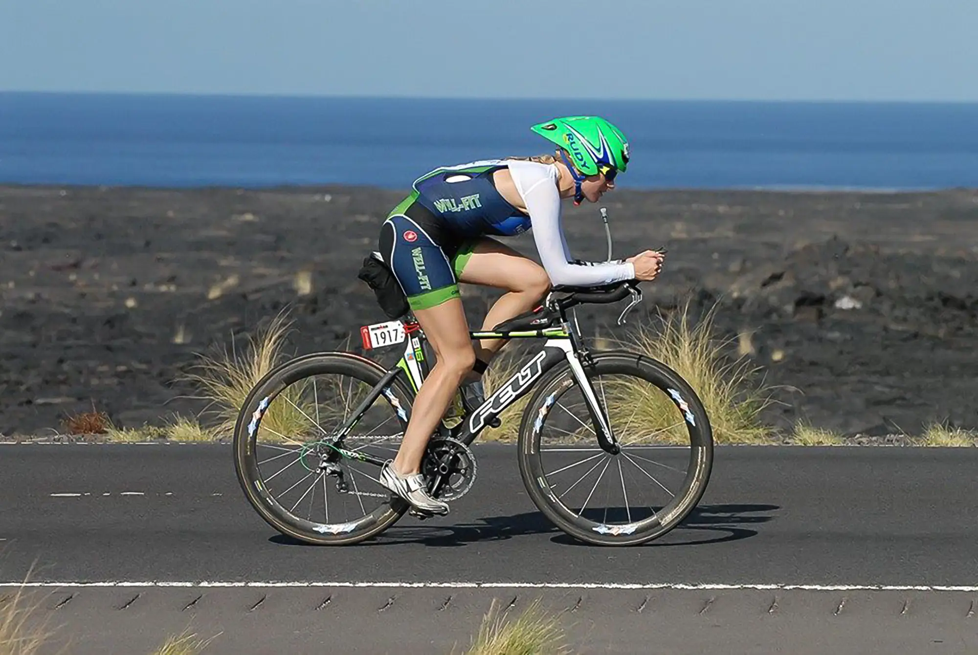Well-Fit athlete riding in aero position on the Kona bike course during the Ironman World Championship, with lava fields and ocean in the background