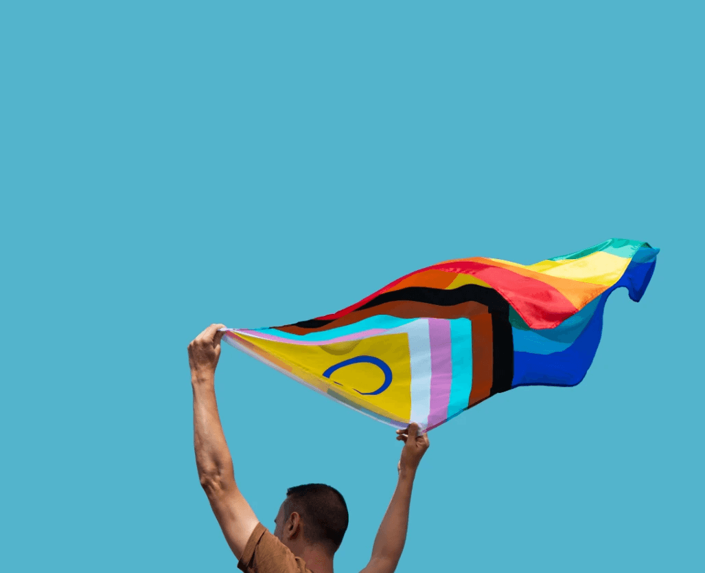 A person holds up a colorful flag against a bright blue sky, celebrating diversity and pride.